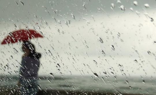pessoa com guarda chuva vista de dentro de uma janela com gotas de chuva e tempo fechado