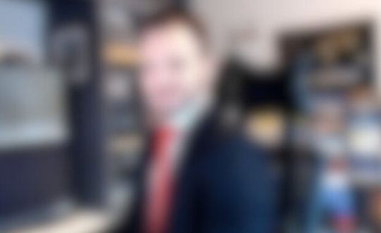 Imagem mostra homem branco de terno e gravata vermelha com uma estante de livros atrás