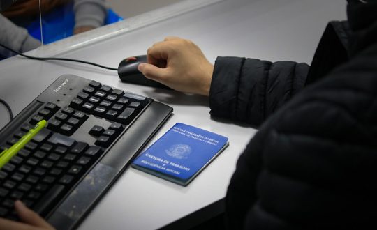 Foto de carteira de trabalho em cima de uma mesa e pessoa mexendo no computador