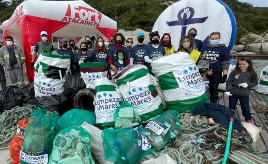 A imagem mostra pessoas em uma praia segurando sacolas cheias de lixo retiradas do mar pela equipe do projeto Limpeza dos Mares, apoiado pelo Fort Atacadista