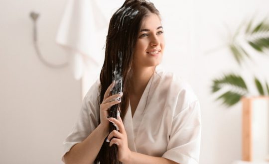Mulher usando um roupão, sentada em um banheiro e passando creme no cabelo