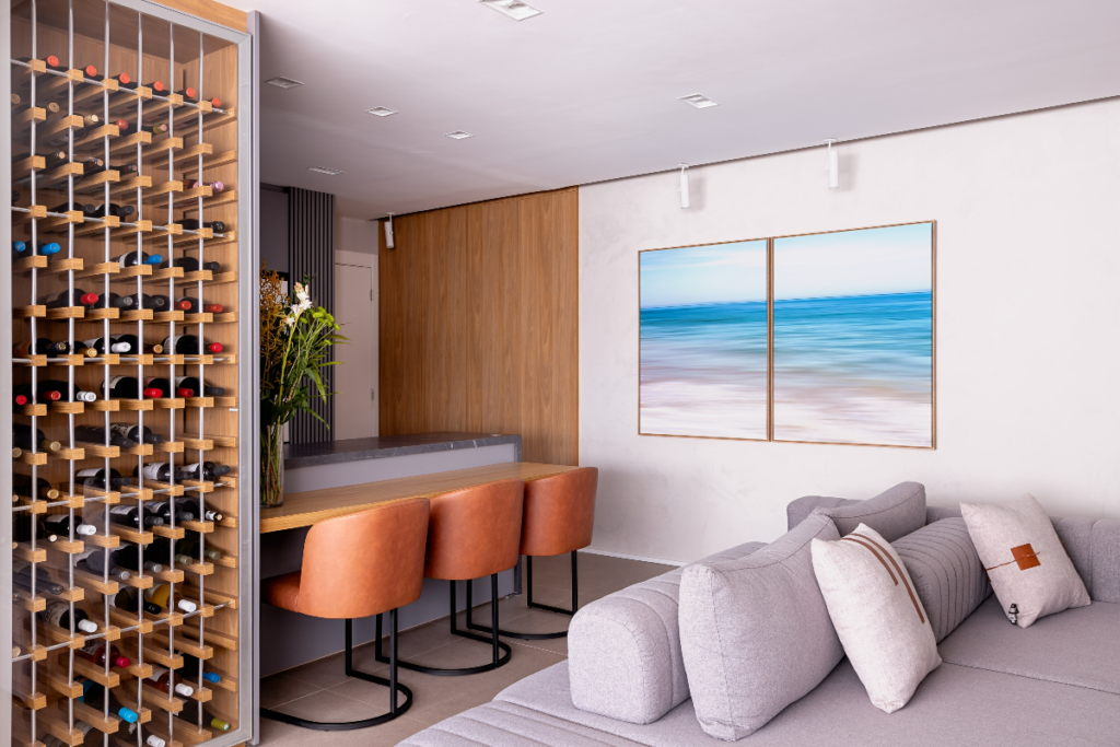 Sala com sofá cinza e almofadas, atrás está a mesa de jantar e ao lado a adega com garrafas de vinho, as cadeiras são laranjas e estofadas e a parede do fundo em cor clara com dois quadros com imagem de praia