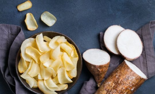 Tigela com chips de mandioca dourados ao lado de raízes cortadas