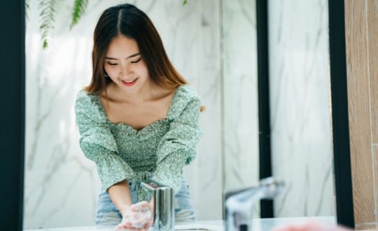 Jovem usando blusa verde com o cabelo liso solto, lavando as mãos no banheiro