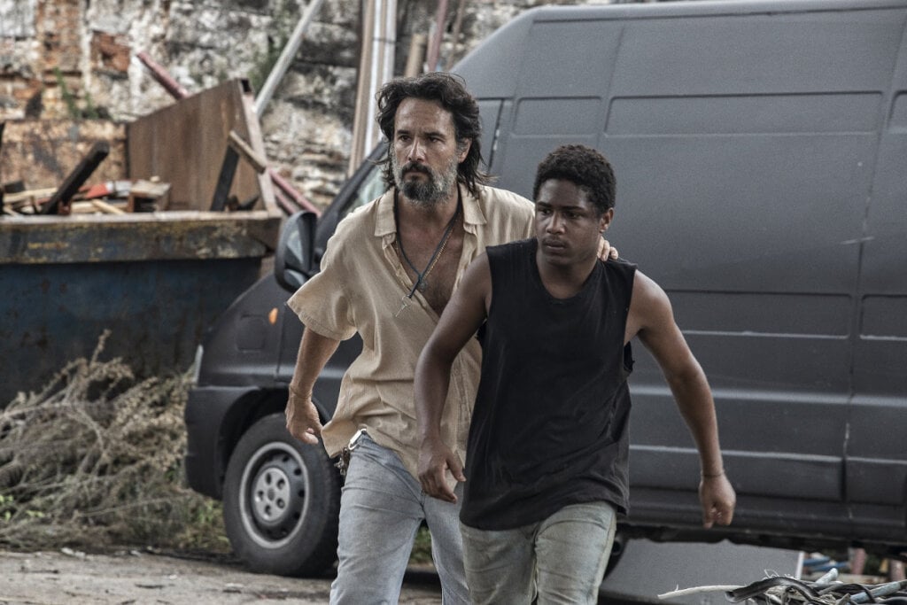 Cena do filme "7 Prisioneiros" com dois homens andando. o jovem negro à frente e homem branco atrás