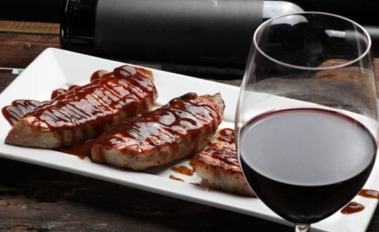 Prato branco com três bifes de porco com molho barbecue ao lado de uma taça com vinho tinto