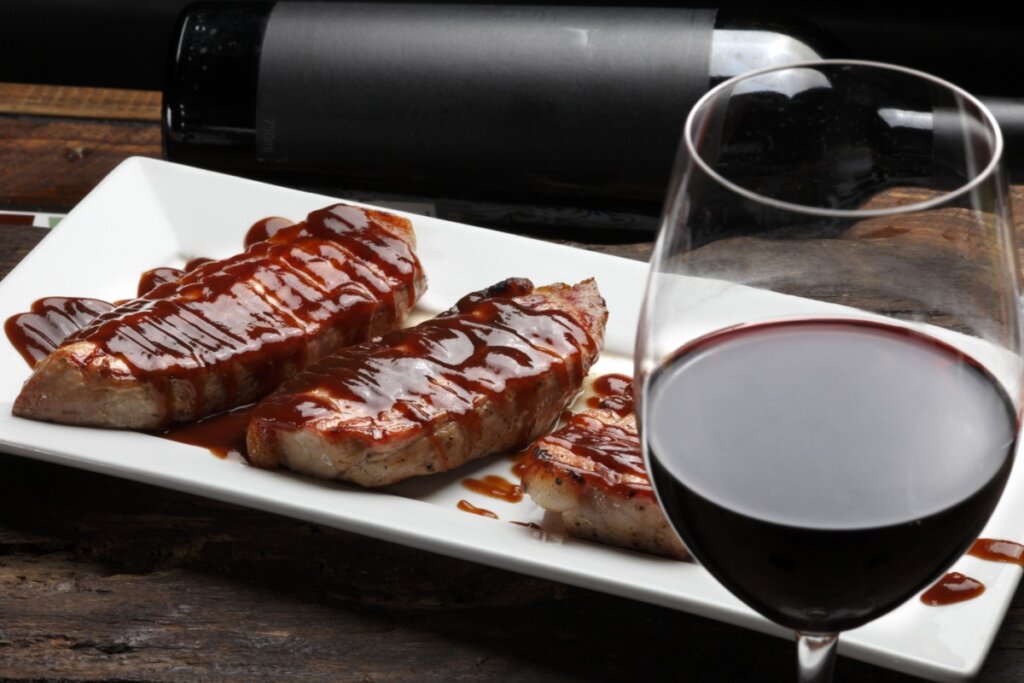 Prato branco com três bifes de porco com molho barbecue ao lado de uma taça com vinho tinto 