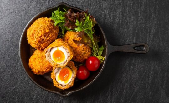 Ovo empanado à moda britânica servido com salada de folhas e tomates-cereja, em uma frigideira de ferro sobre fundo escuro