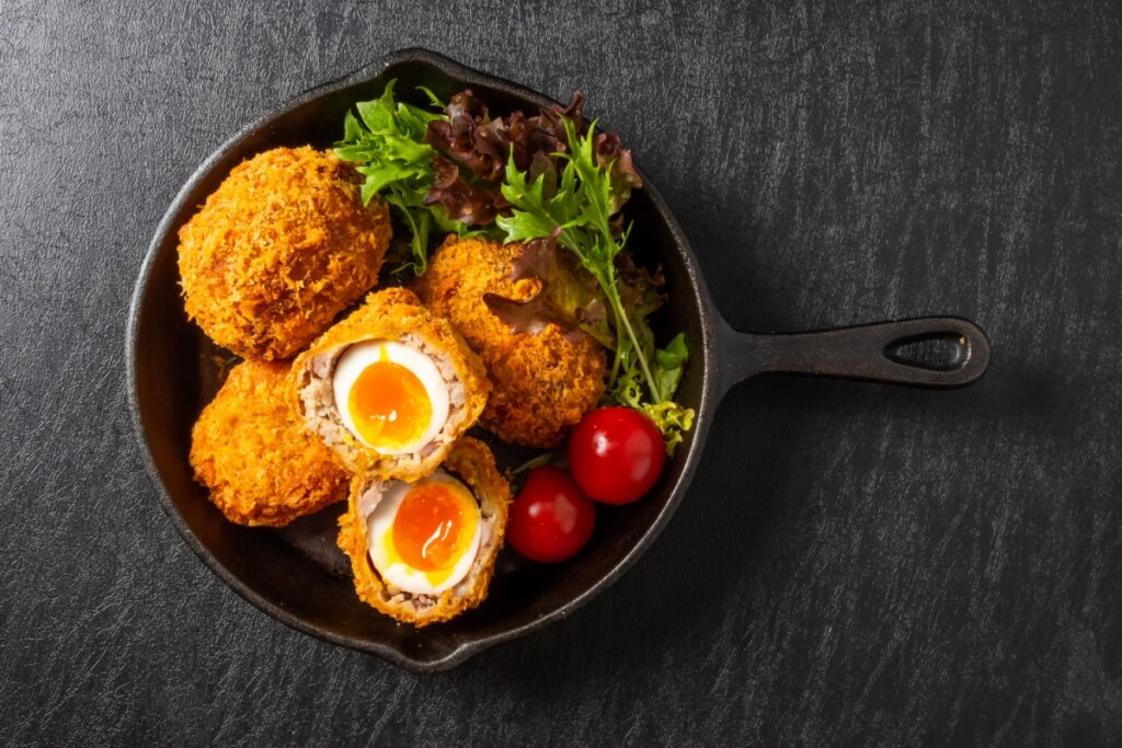 Ovo empanado à moda britânica servido com salada de folhas e tomates-cereja, em uma frigideira de ferro sobre fundo escuro