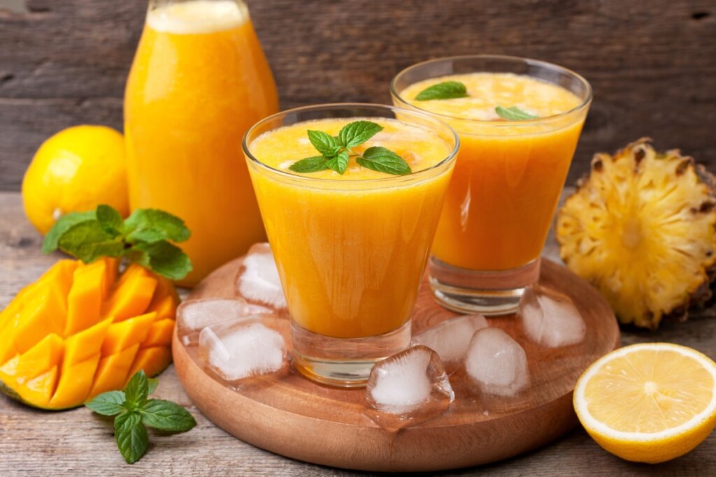 Dois copos de vidro com suco de manga, abacaxi e hortelã em descanso de madeira ao lado de manga, abacaxi, laranja cortados e folhas de hortelã