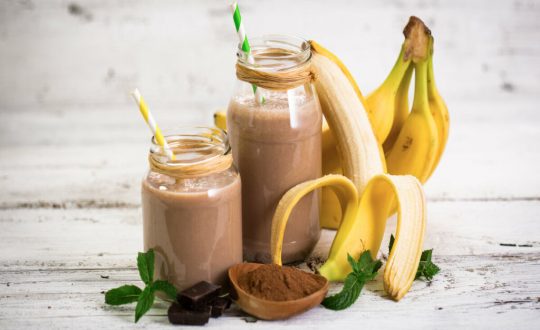 Dois copos com vitamina de banana e chocolate em cima de uma mesa de madeira com bananas e chocolate em pó