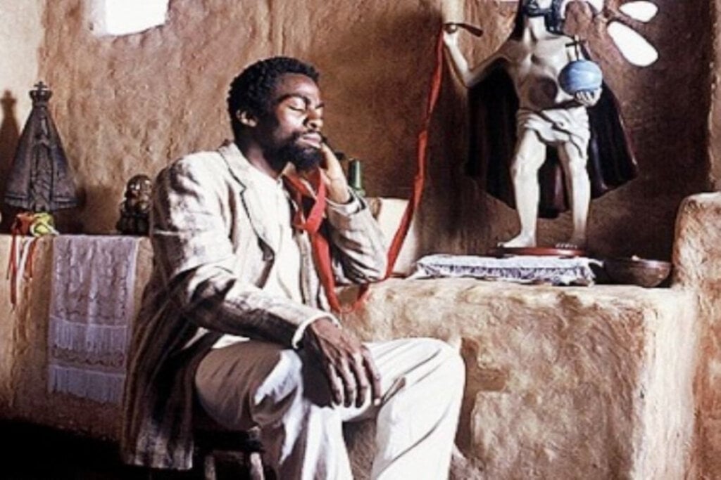 Cena do filme "Cafundó" com homem negro sentado, com os olhos fechados e cotovelo em mesa de pedra