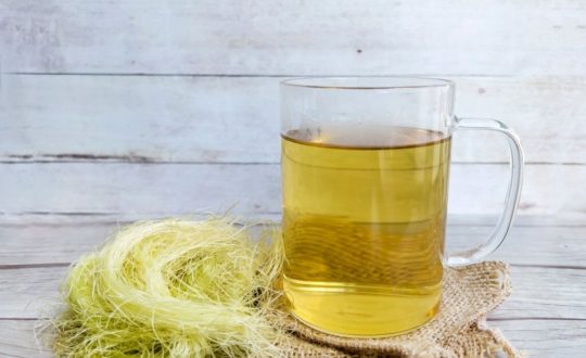 Copo de chá de cabelo de milho ao lado de fios secos da planta sobre uma superfície de madeira clara