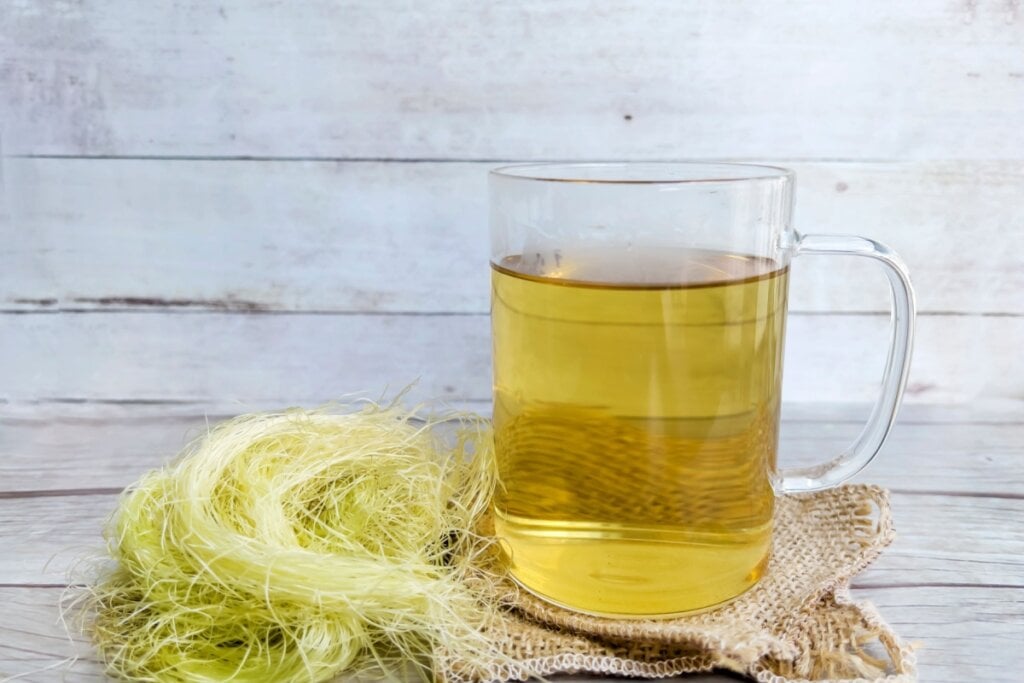 Copo de chá de cabelo de milho ao lado de fios secos da planta sobre uma superfície de madeira clara