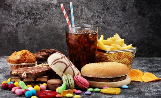 Alimentos processados e ricos em açúcar e gordura em cima de mesa de pedra como hambúrguer, refrigerante, chocolate e batata frita