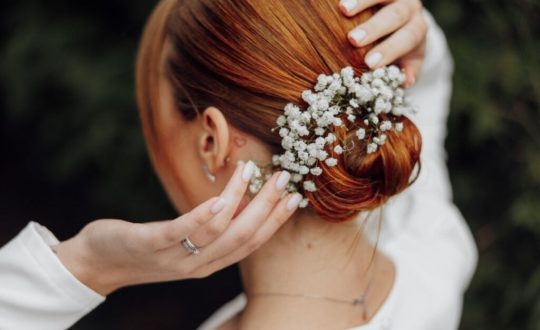 Mulher com cabelo ruivo preso em coque baixo com um arranjo de flores vestida de noiva e com as mãos no cabelo