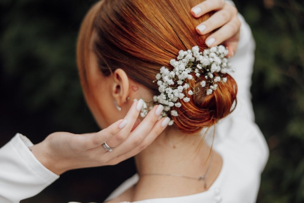 Mulher com cabelo ruivo preso em coque baixo com um arranjo de flores vestida de noiva e com as mãos no cabelo 