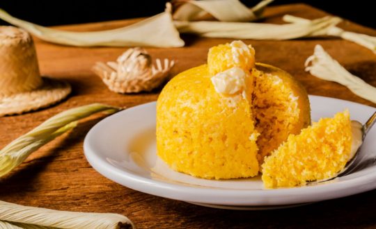 Cuscuz com manteiga em prato branco. Ao fundo palhas de milho