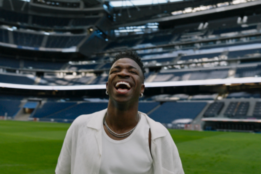 Homem negro com o cabelo curto, e usando regata e camisa branca sorrindo, no meio do gramado de estádio de futebol