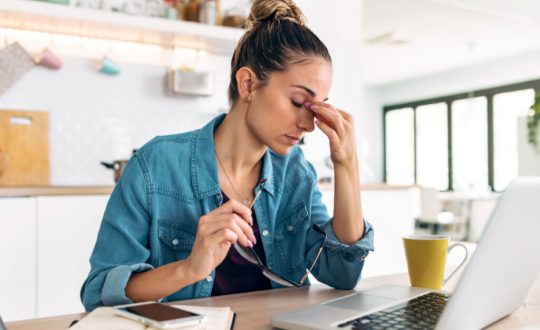 Mulher de camisa jeans, sentada, com uma mão nos olhos e outra segurando os óculos. notebook em cima de mesa à frente dela