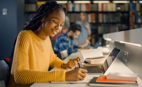 Estudante com o cabelo trançado, preso, usando um suéter amarelo escrevendo na biblioteca da universidade