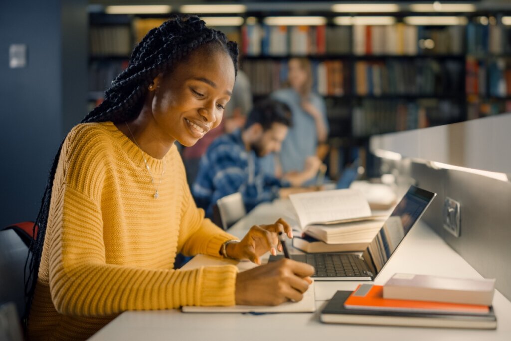 Estudante com o cabelo trançado, preso, usando um suéter amarelo escrevendo na biblioteca da universidade