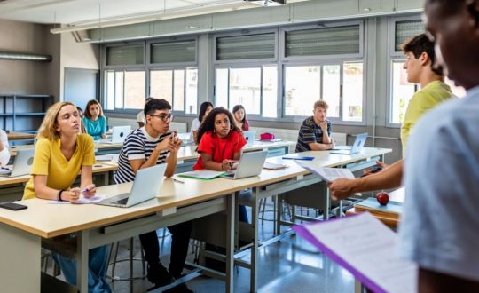 Alunos sentados em mesas na sala de aula assistindo apresentação de projeto de um colega que está a frente da sala