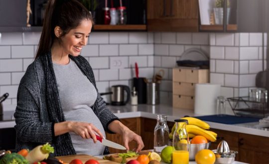 Mulher grávida com o cabelo preto amarrado, usando uma camiseta cinza e um casaco preto cortando frutas em bancada na cozinha