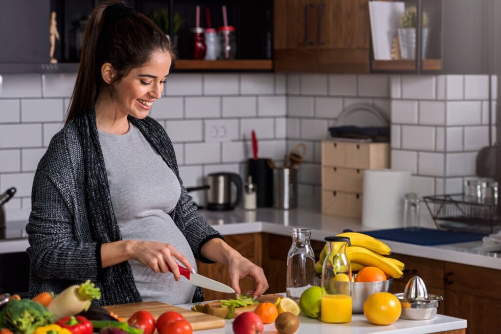 Mulher grávida com o cabelo preto amarrado, usando uma camiseta cinza e um casaco preto cortando frutas em bancada na cozinha