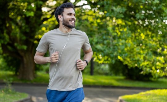 Homem com cabelo curto, barba e usando camisa cinza e short azul correndo no parque com fones de ouvido
