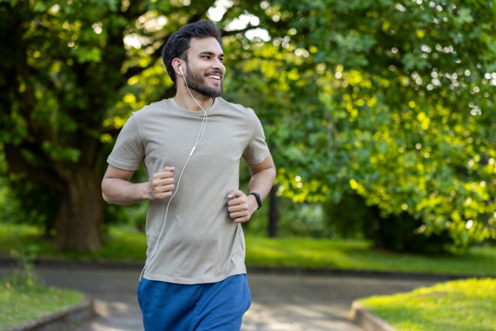 Homem com cabelo curto, barba e usando camisa cinza e short azul correndo no parque com fones de ouvido