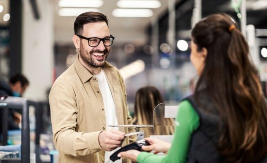 Homem com cabelo curto e barba, usando óculos preto. Uma camiseta branca e uma camisa bege fazendo o pagamento de uma compra