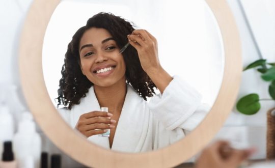Jovem negra com o cabelo cacheado usando roupão branco e passando sérum no rosto em frente ao espelho