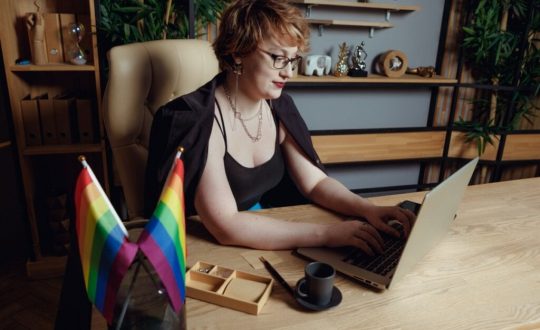 Mulher de cabelos curtos ruivos mexendo em notebook em sua sala de trabalho, em que há duas bandeiras LGBT
