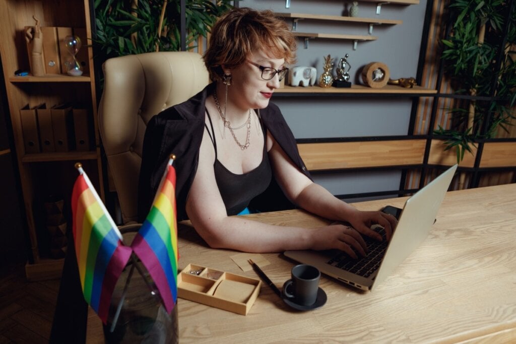 Mulher de cabelos curtos ruivos mexendo em notebook em sua sala de trabalho, em que há duas bandeiras LGBT