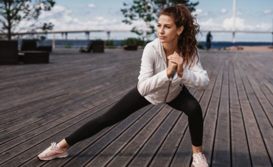 mulher de calça preta e blusa branca. ela está fazendo exercícios em chão de madeira. uma perna está esticada e a outra flexionada