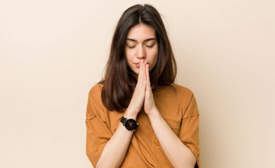 Mulher com camiseta marrom, olhos fechados e rezando