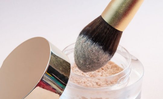 Pincel de pó dourado sendo aplicado em vidro de maquiagem em fundo branco