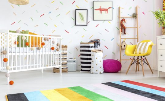 Quarto de bebê com cadeira amarela, berço branco, tapete e decorações muito coloridas.