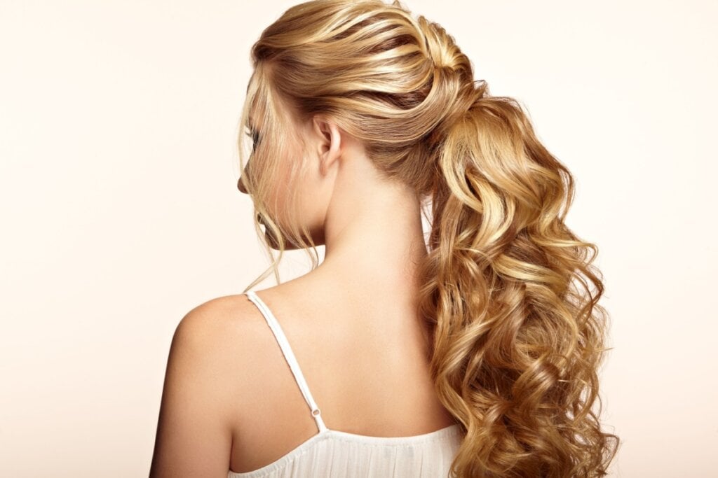 Mulher com cabelo loiro cacheado preso em rabo de cavalo volumoso usando um vestido branco de alcinhas 