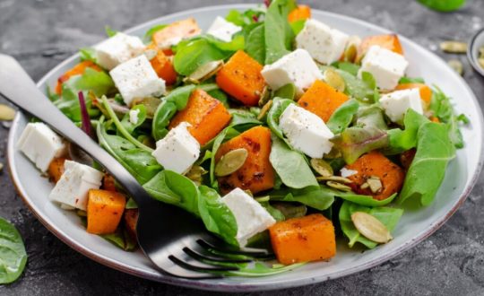 Salada de abóbora com rúcula e queijo feta servida em prato grande branco em cima de mesa de pedra