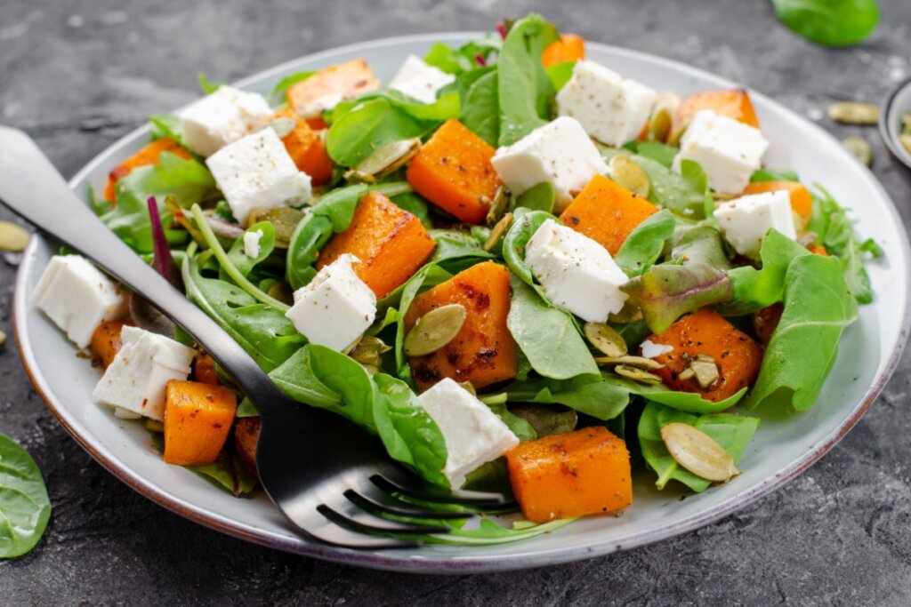 Salada de abóbora com rúcula e queijo feta servida em prato grande branco em cima de mesa de pedra