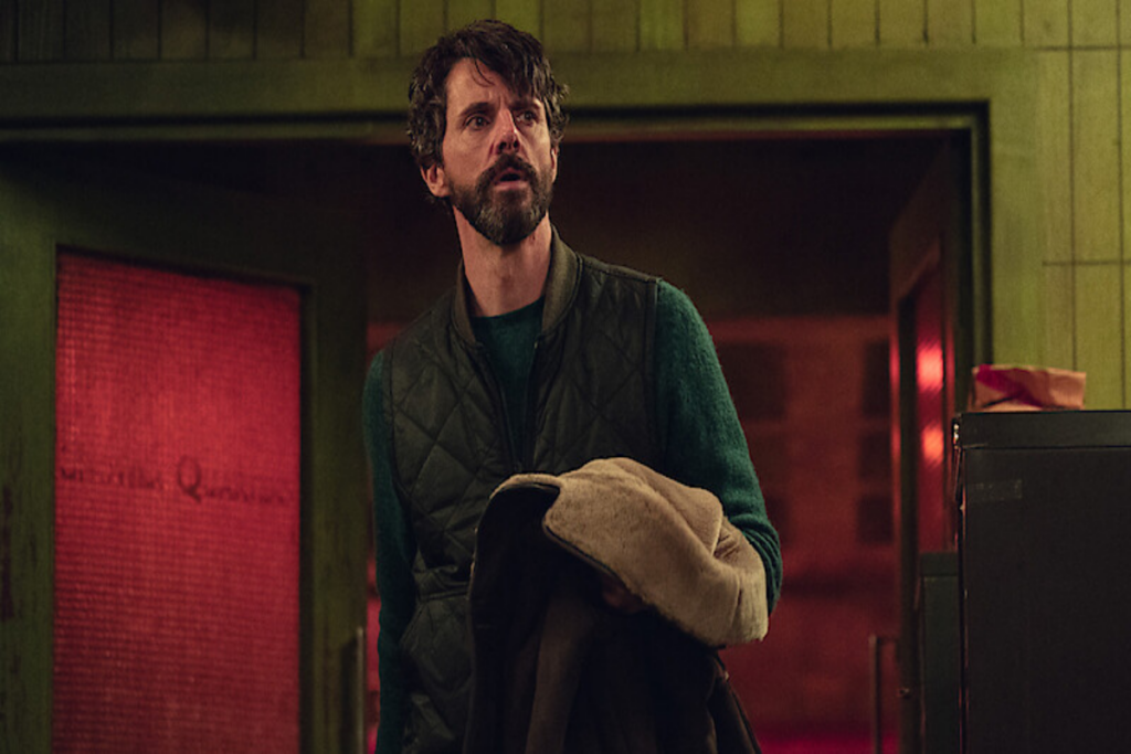 Homem com cabelo curto castanho, de barba usando uma camisa manga longa verde e um colete verde escuro segurando um casaco marrom e bege