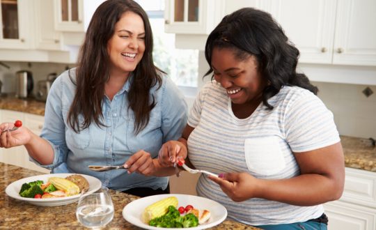 Duas mulheres sorrindo comendo prato com alimentos saudáveis em uma mesa