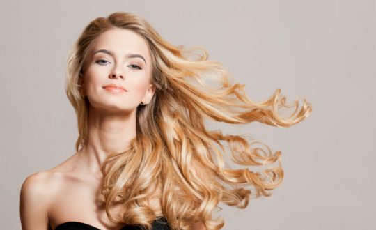 Mulher com cabelo loiro claro ondulado comprido e usando top preto em fundo bege