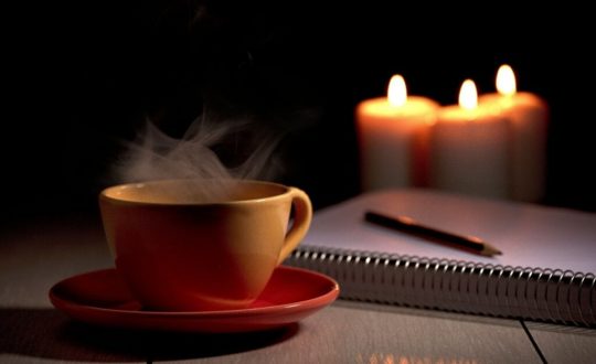 Xícara de café amarela ao lado de caderno e caneta e três velas ao fundo