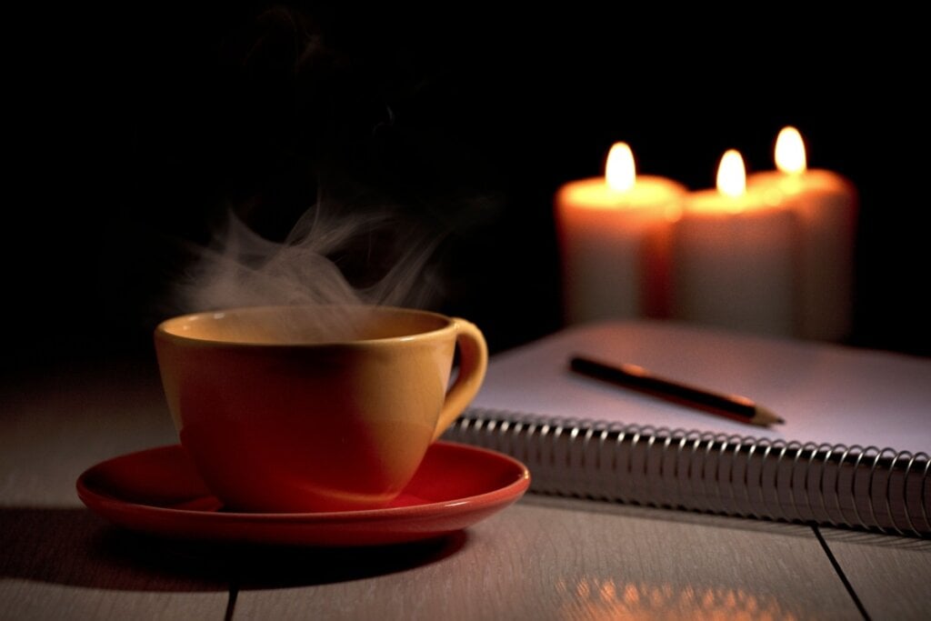 Xícara de café amarela ao lado de caderno e caneta e três velas ao fundo