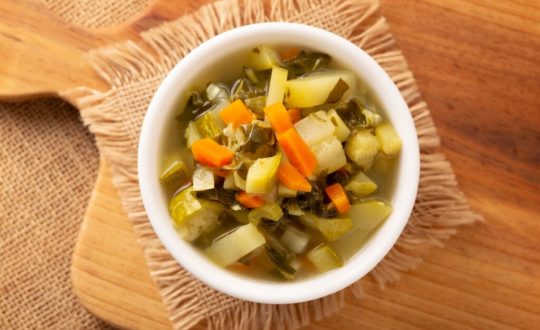 Sopa de legumes servido em tigela branca em cima de tabua de madeira em cima de mesa de madeira