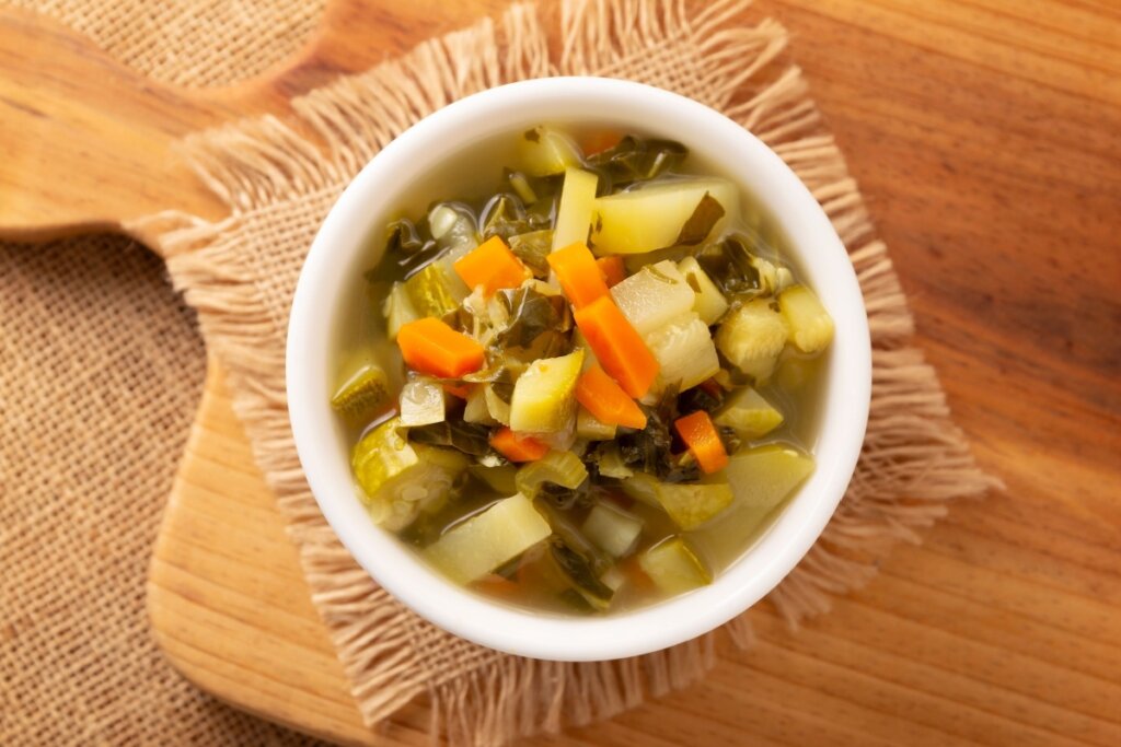 Sopa de legumes servido em tigela branca em cima de tabua de madeira em cima de mesa de madeira