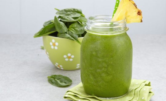 Suco verde em copo de vidro com canudo verde e fatia de abacaxi. Pote verde com espinafre ao fundo